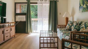 A room with a wooden floor, a sofa with floral upholstery, a wooden chair, a small table, a television, an air conditioning unit, and a window with a view of greenery.