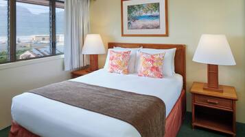 A hotel room with a large bed, two bedside tables with lamps, a painting on the wall, and a window with a view of the beach and mountains.