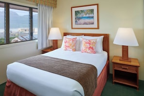 A hotel room with a large bed, two bedside tables with lamps, a painting on the wall, and a window with a view of the beach and mountains. - Banyan Harbor Resort (Lihue)