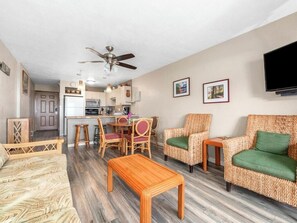 A living room with a sofa, a wooden coffee table, wicker chairs, a ceiling fan, a kitchen area, and a flat-screen TV mounted on the wall. - Banyan Harbor Resort (Lihue)