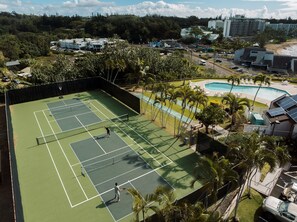 Vista aèria d'una pista de tennis envoltada de palmeres i una zona de piscina.
