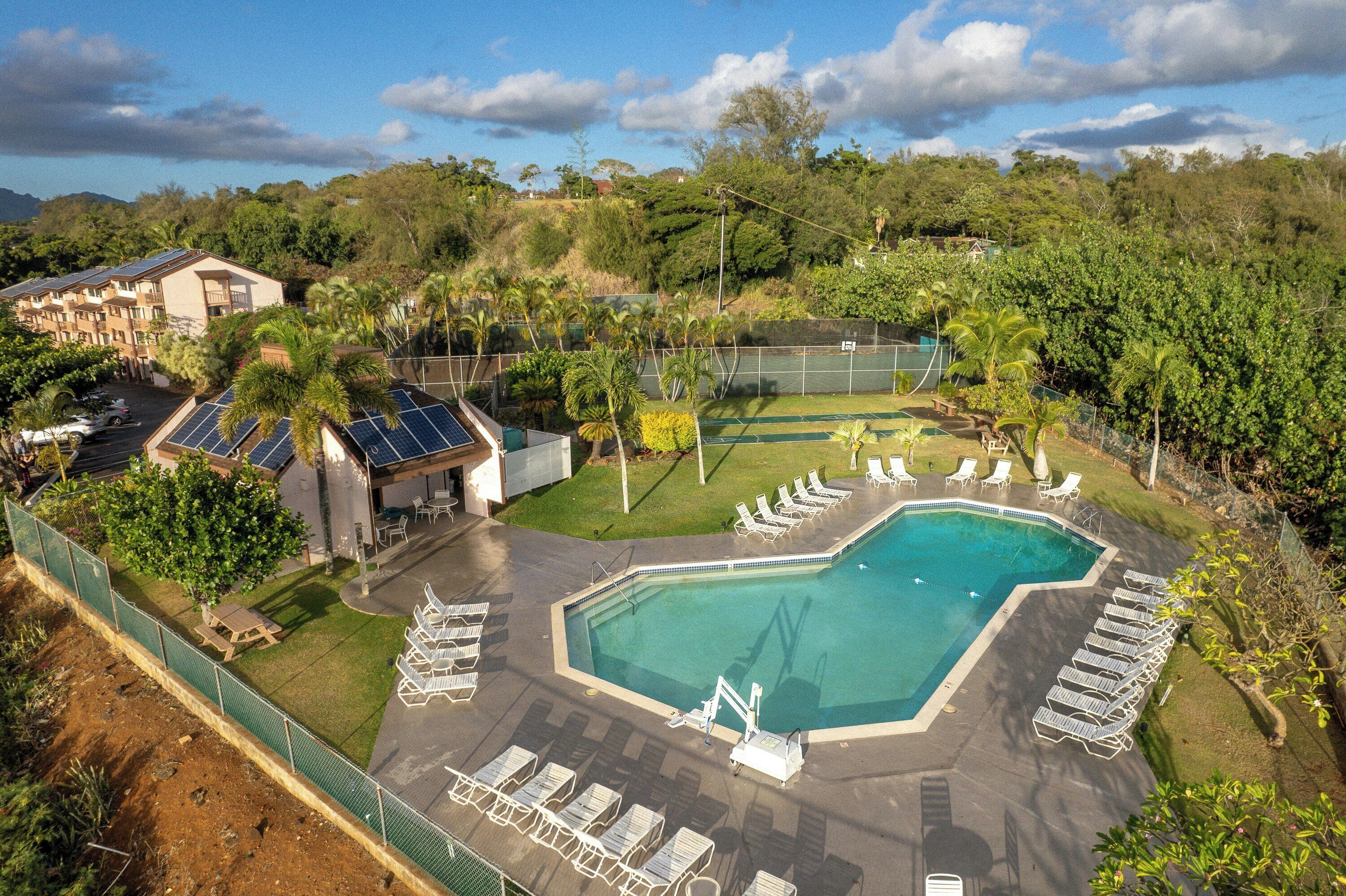A swimming pool with sun loungers, a tennis court, and a building with solar panels.