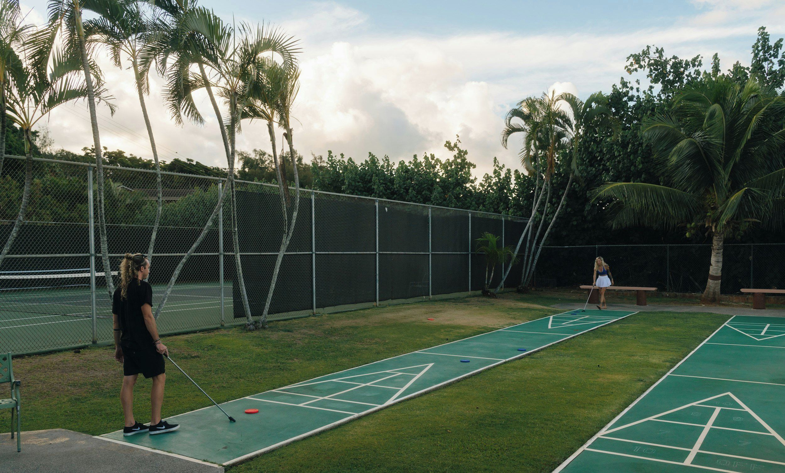 A shuffleboard court with a chain-link fence and palm trees in the background.