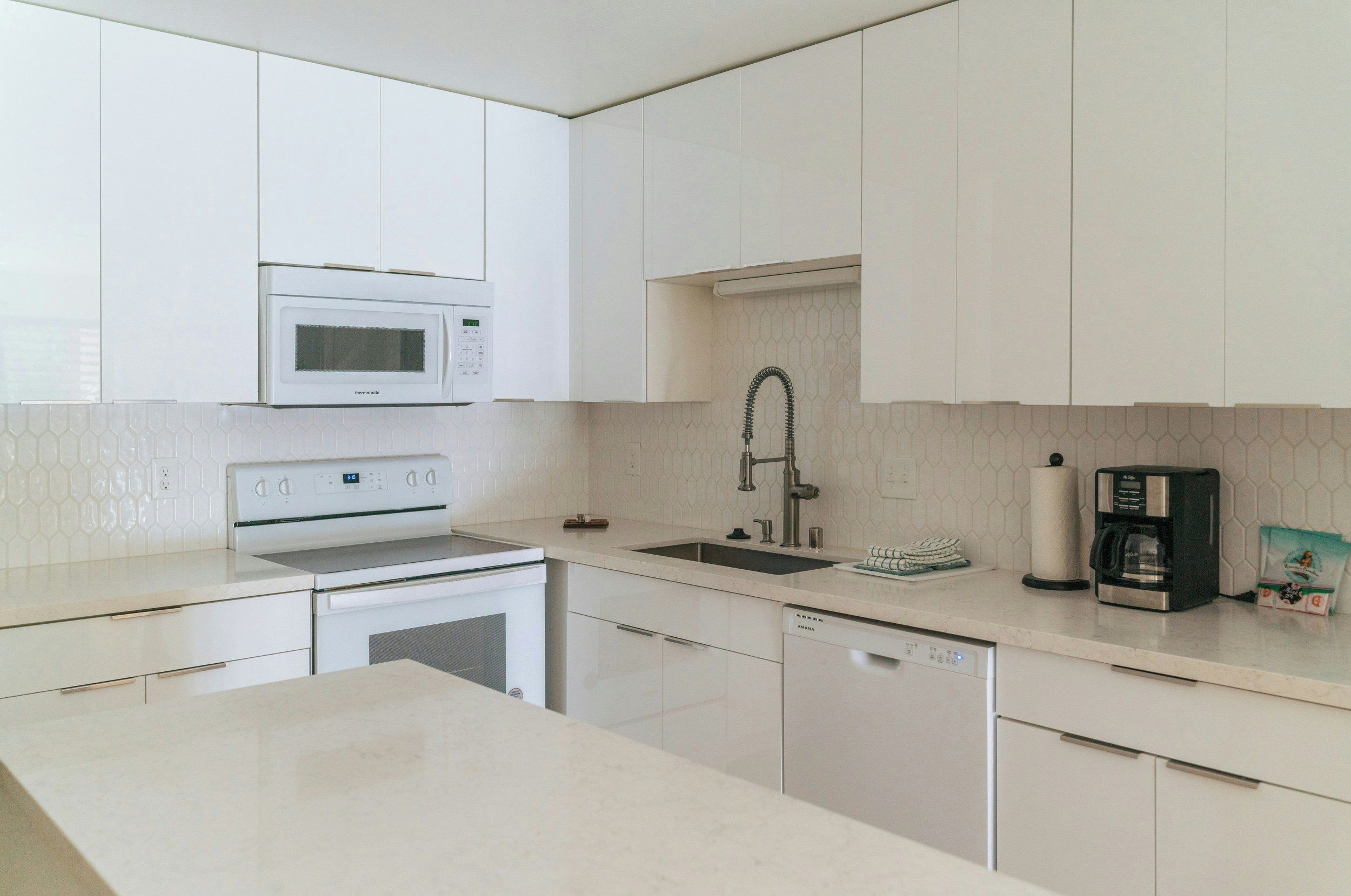 Una cocina moderna con armarios blancos, microondas integrado, pileta de acero inoxidable y cafetera.