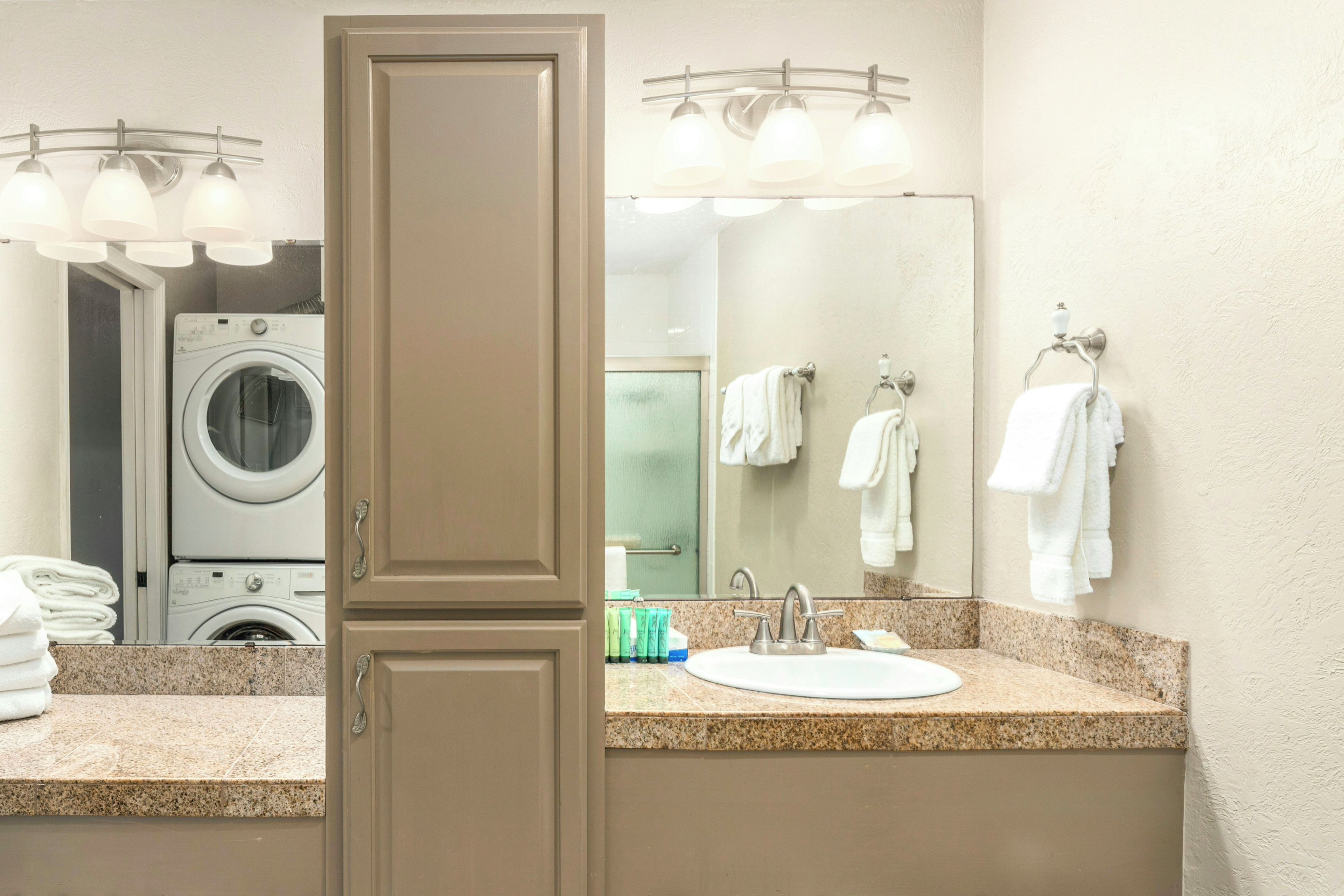 A hotel bathroom with a sink, mirror, towel rack, and a washing machine in the background.