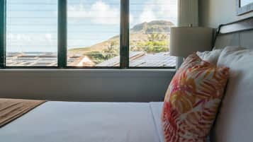 Quarto de hotel com uma cama, janela com vista para o oceano e uma montanha ao longe, e um abajur na mesa de cabeceira.