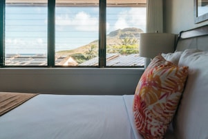 Quarto de hotel com uma cama, janela com vista para o oceano e uma montanha ao longe, e um abajur na mesa de cabeceira.