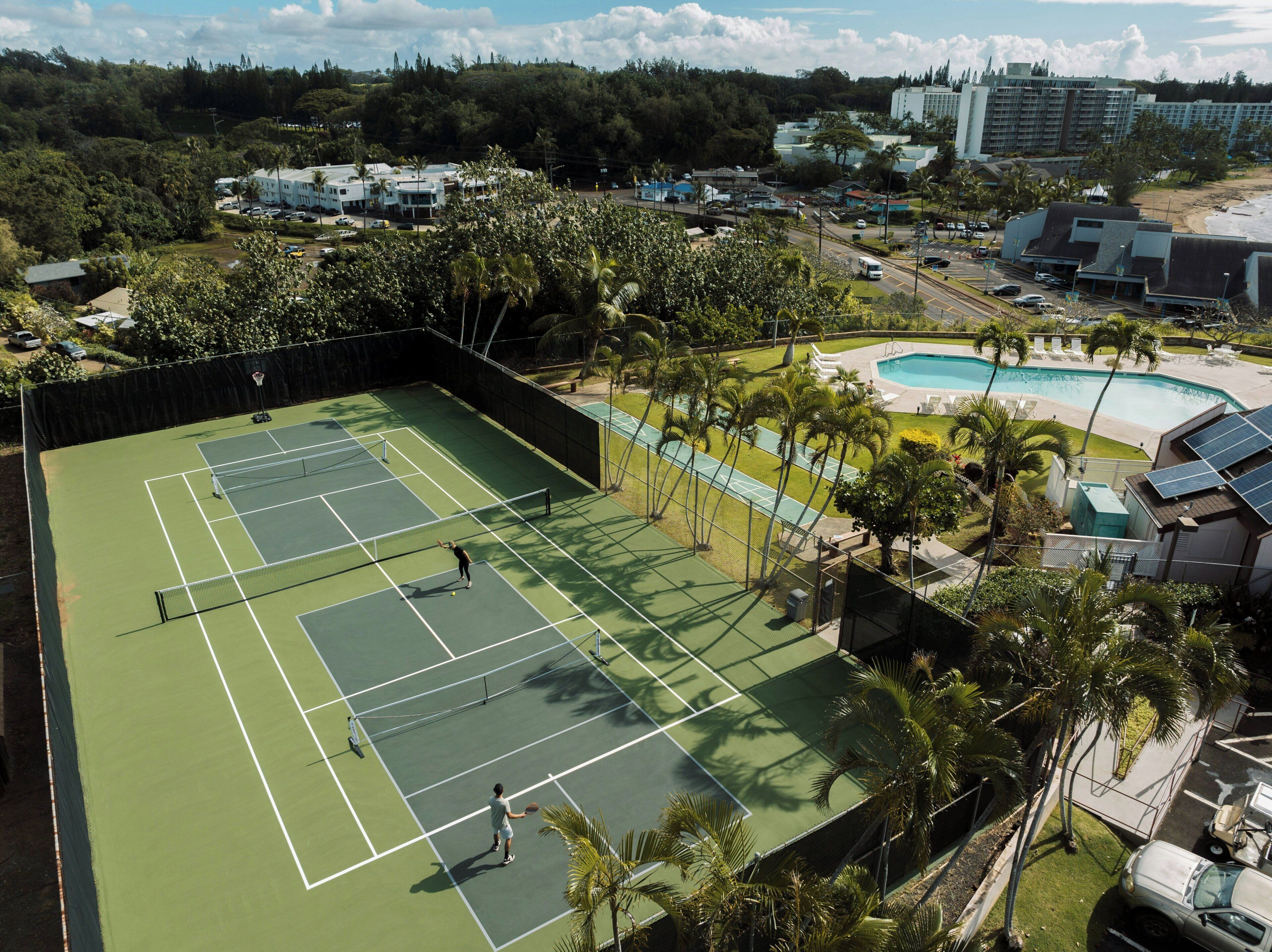 A tennis court surrounded by palm trees and a swimming pool.