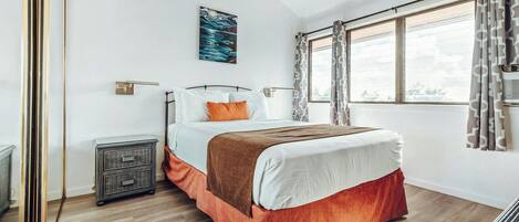 Une chambre à coucher comprenant un lit, une table de chevet, une lampe fixée au mur, une fenêtre avec des rideaux et un ventilateur de plafond.
