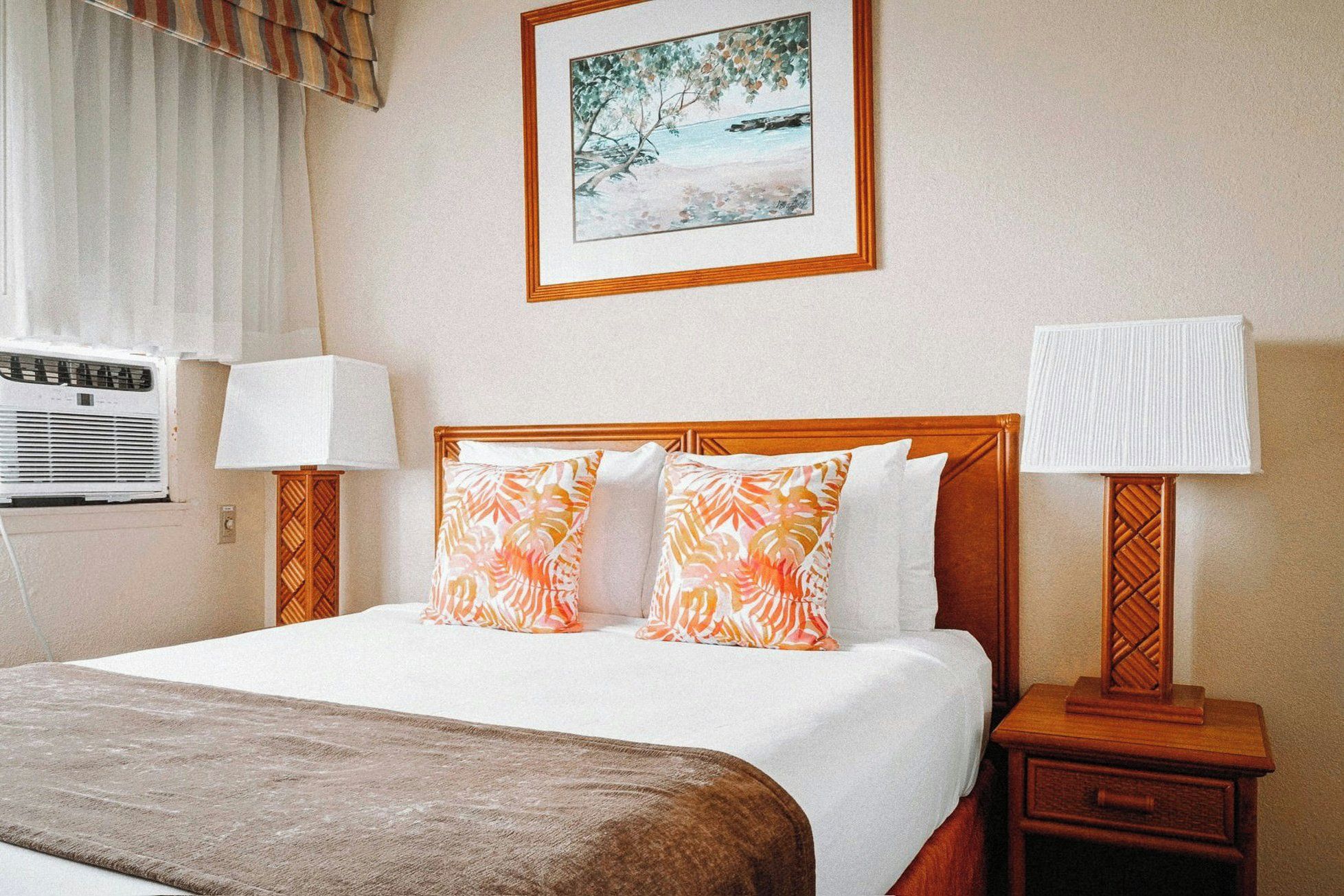A hotel room with a bed, two pillows, a bedside table with a lamp, a wall-mounted air conditioner, and a framed picture above the bed.