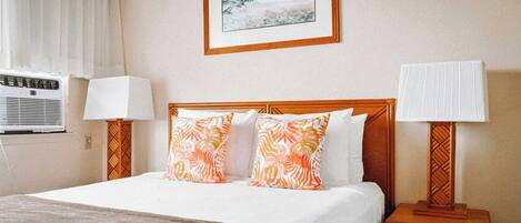 A hotel room with a bed, two pillows, a bedside table with a lamp, a wall-mounted air conditioner, and a framed picture above the bed.