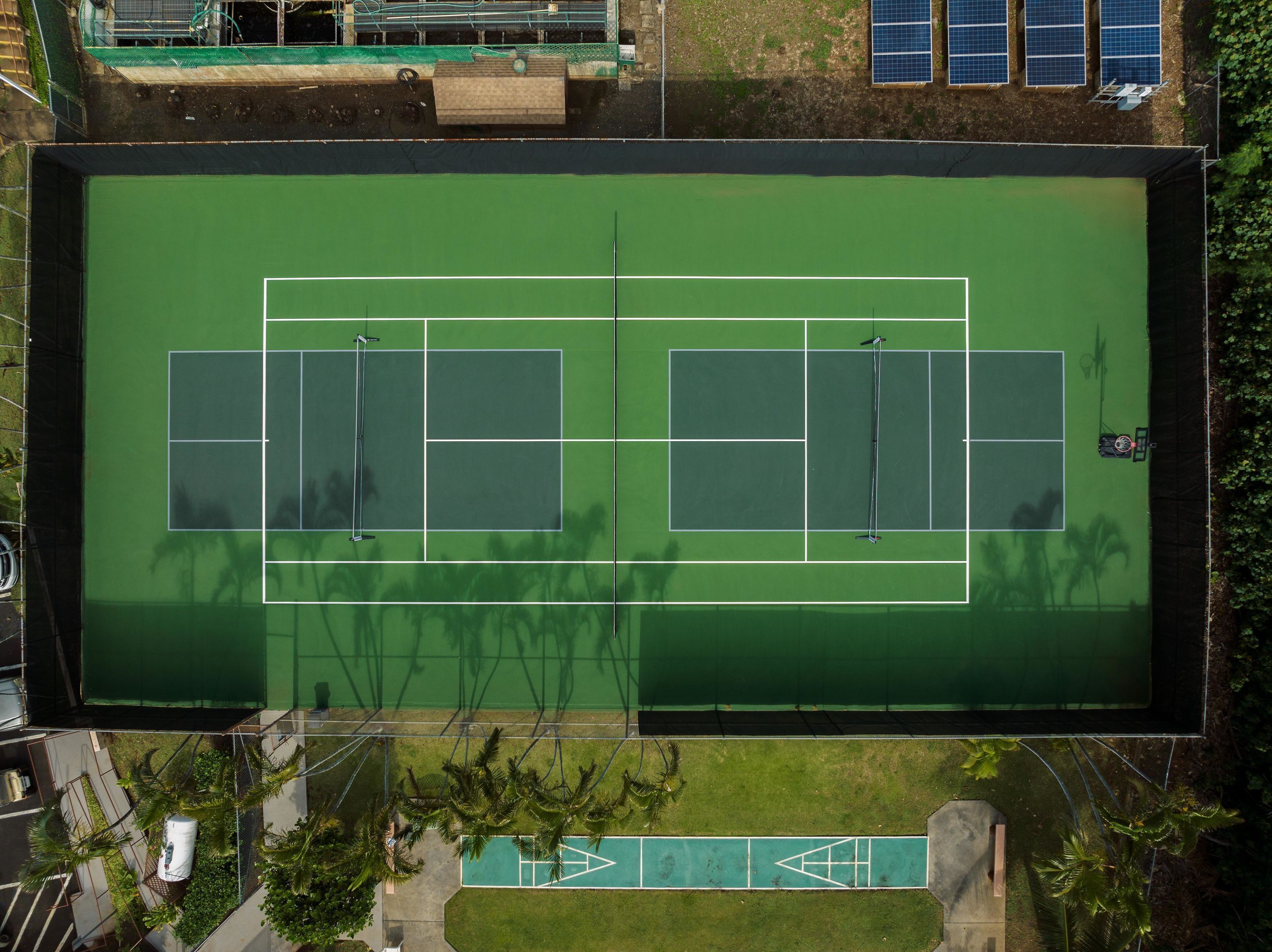 An aerial view of a tennis court with two courts, surrounded by greenery and solar panels.
