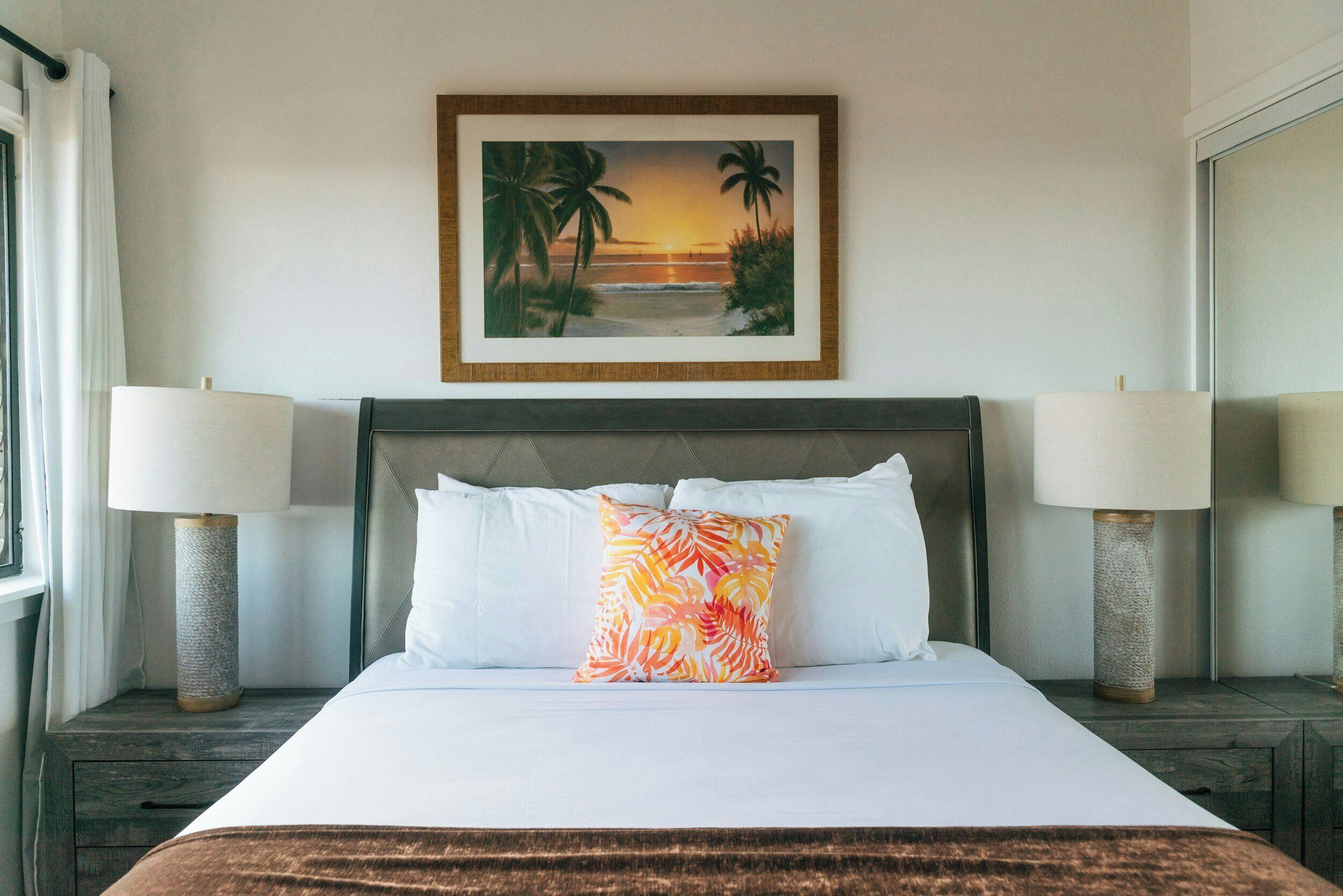A hotel room with a bed, two bedside lamps, a framed picture of a beach scene, and a brown throw blanket.