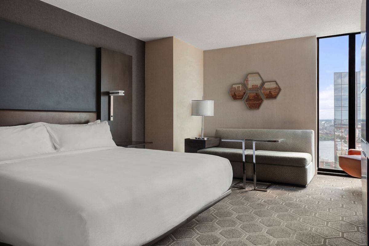 A modern hotel room with a large bed, a sofa, a desk, and a view of the city through large windows.