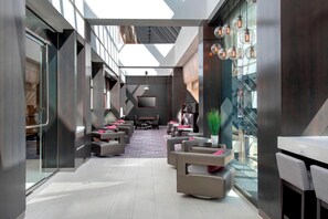 Eine moderne Hotellobby mit hoher Decke, Glaswänden und einer Sitzecke mit Sofas und Sesseln.