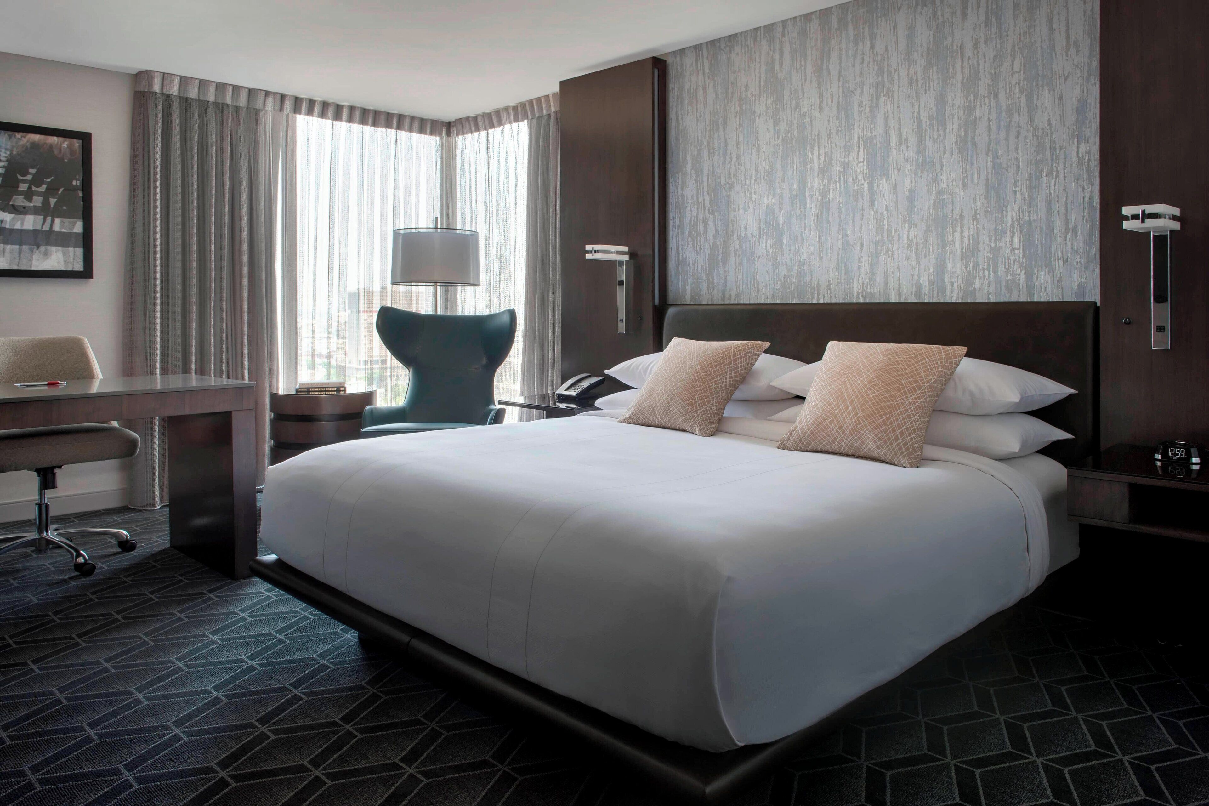 Habitación de hotel con una cama grande, un escritorio, una silla, una lámpara y vistas a la ciudad a través de la ventana.