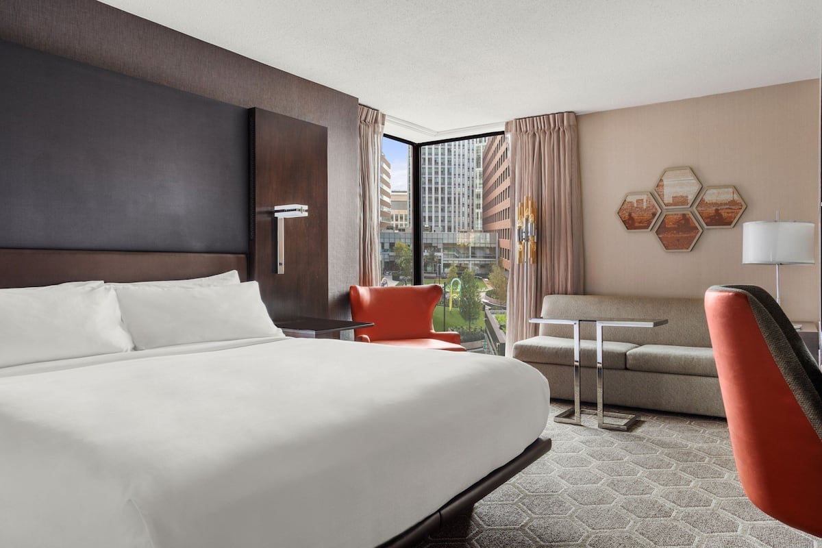 A hotel room with a large bed, a sofa, a table, and a chair. There is a view of buildings through the window.