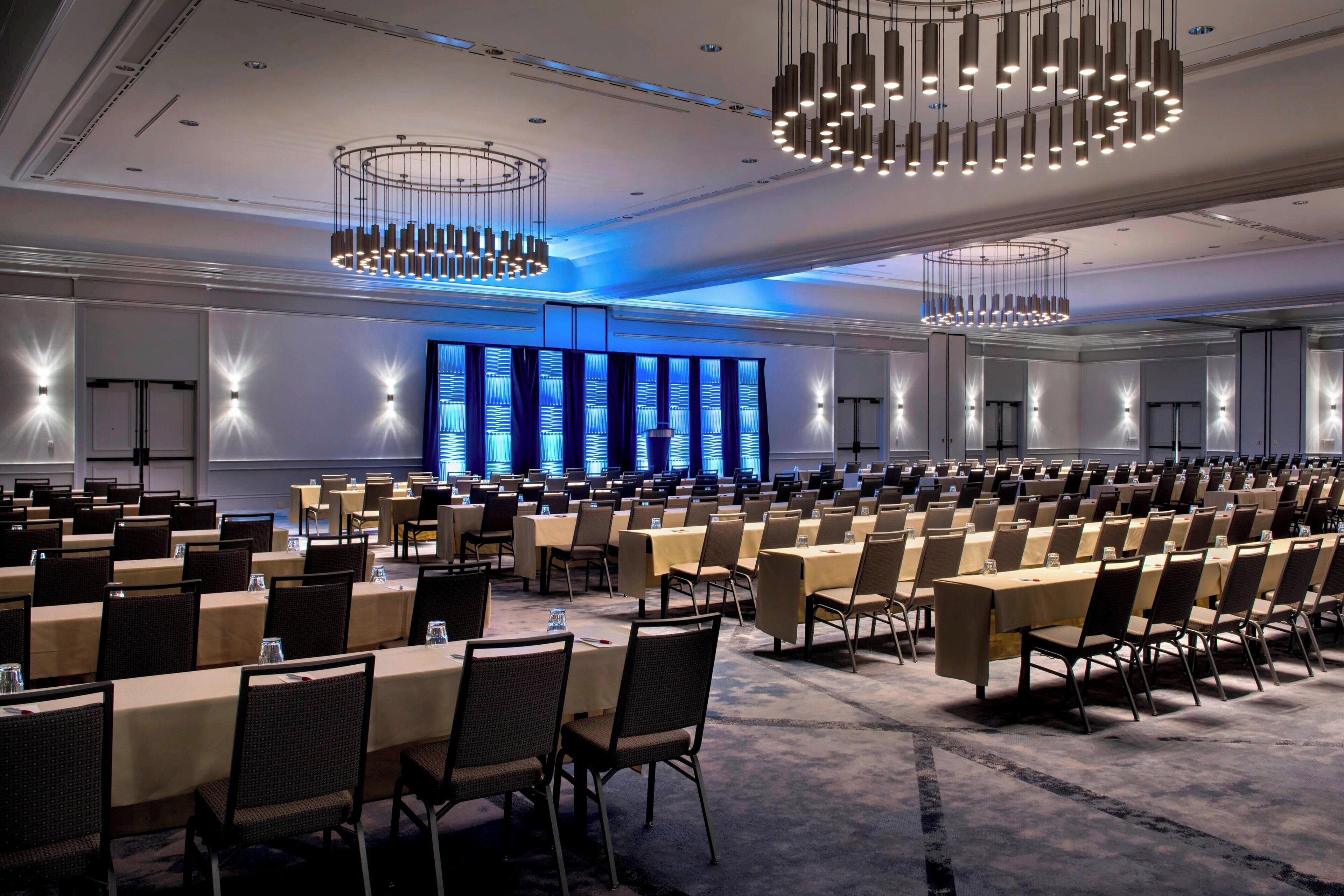 A large conference room with rows of tables and chairs, modern chandeliers, and a blue-lit backdrop.