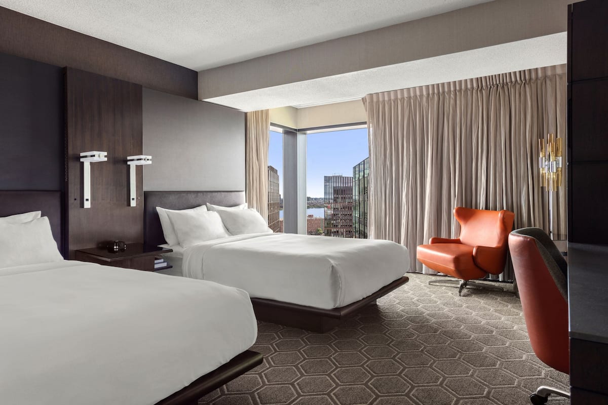 A hotel room with two beds, a large window with curtains, a city view, and a modern orange chair.