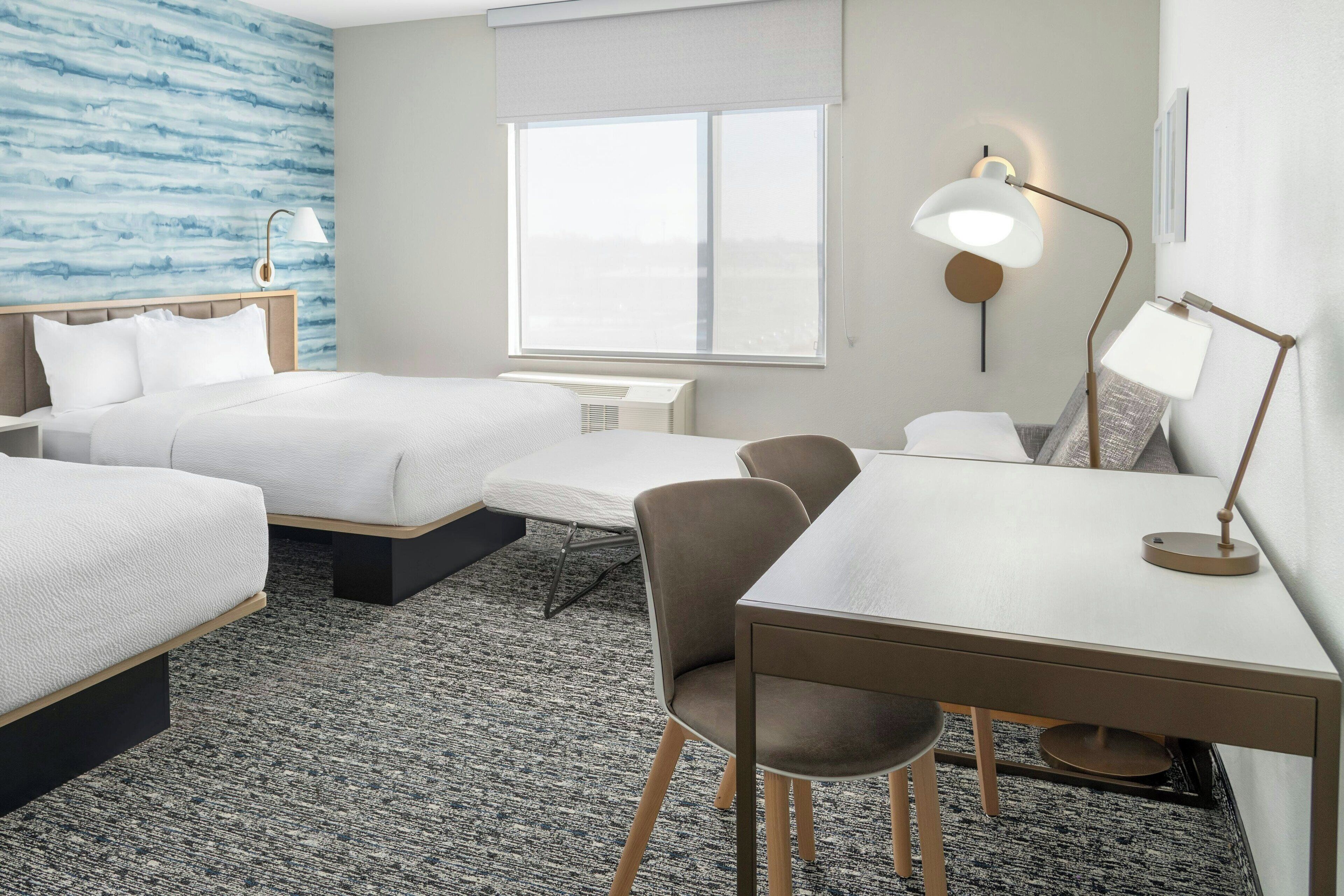 A hotel room with two beds, a desk, a chair, a lamp, and a large window.