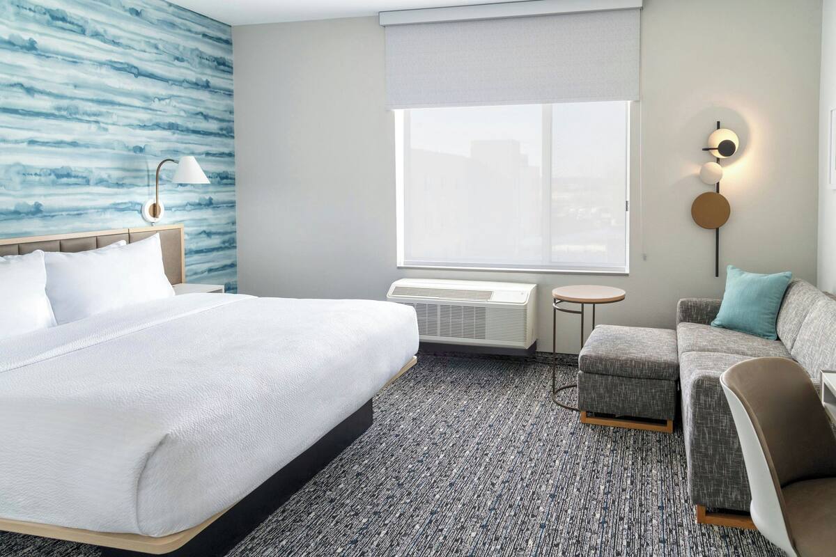 Ein Hotelzimmer mit einem großen Bett, einem grauen Sofa, einem kleinen runden Tisch und einer Wandlampe.