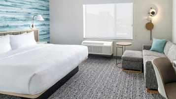 Habitación de hotel con una cama grande, un sofá gris, una mesita redonda y una lámpara de pared.