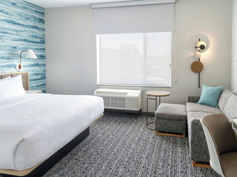 Habitación de hotel con una cama grande, un sofá gris, una mesita redonda y una lámpara de pared.