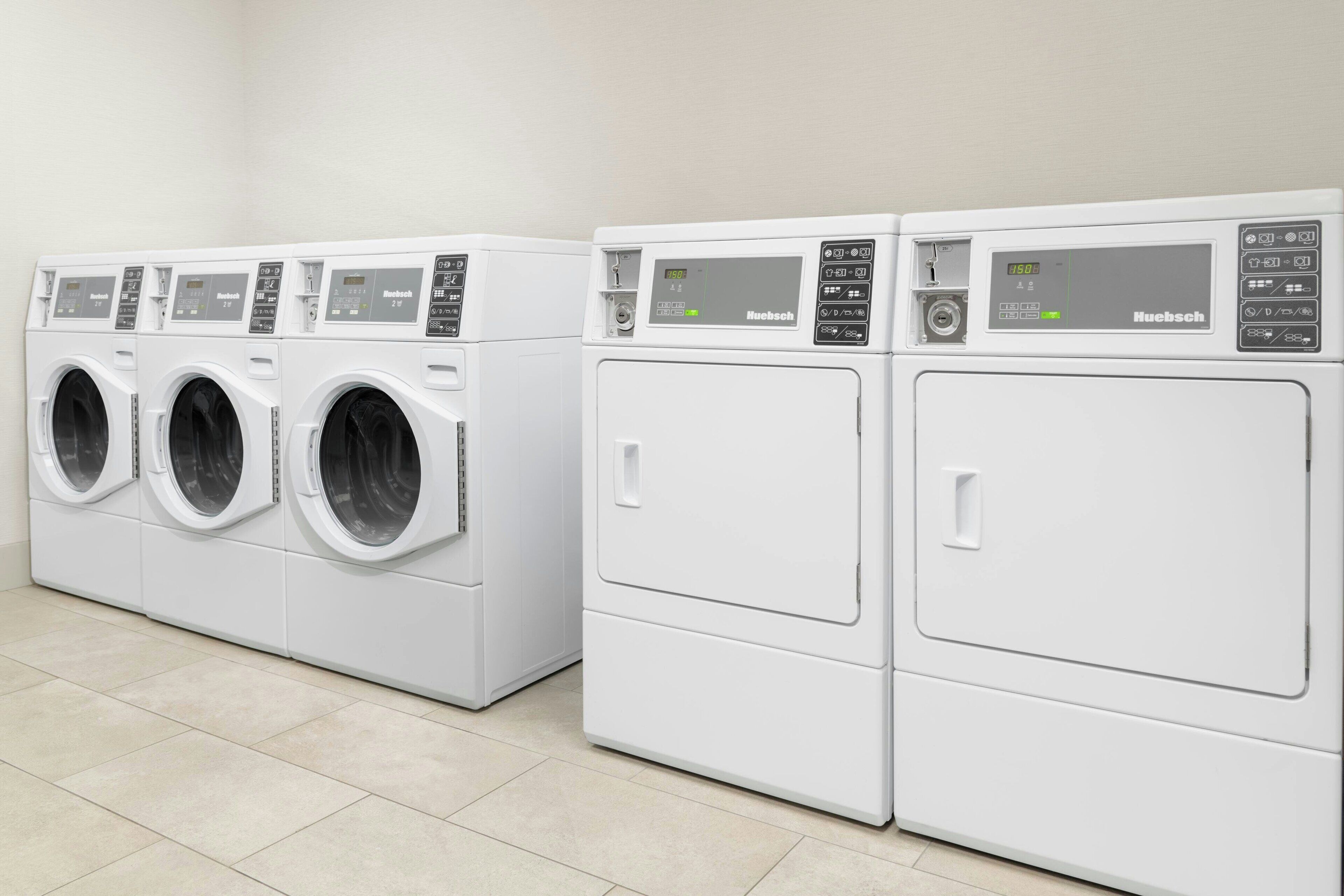 A row of white, modern, front-loading washing machines with digital displays, in a clean, tiled laundry room.