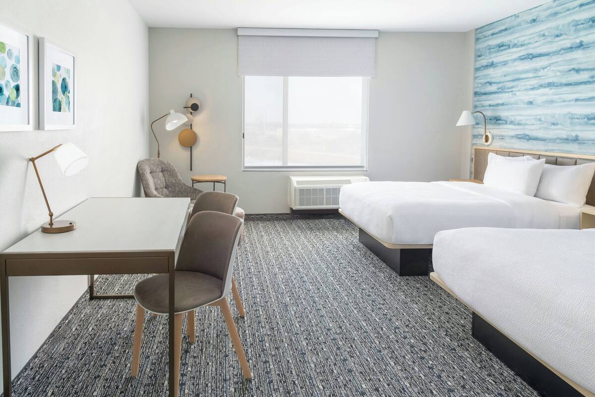 Ein Hotelzimmer mit Schreibtisch, zwei Stühlen, einem Bett, einem Fenster mit Blick und einer Wand mit Kunstwerken.