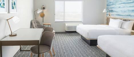 A hotel room with a desk, two chairs, a bed, a window with a view, and a wall with artwork.