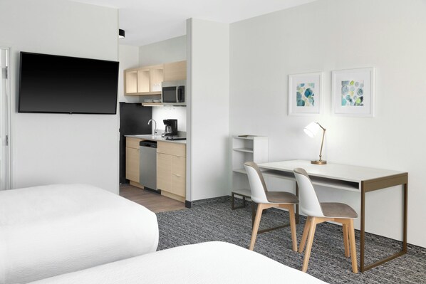 Habitación de hotel con cama, escritorio con dos sillas, televisor, kitchenette y obras de arte en la pared.