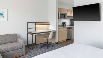 Habitación de hotel moderna con cama, sofá gris, escritorio con silla, televisor de pantalla plana y kitchenette.