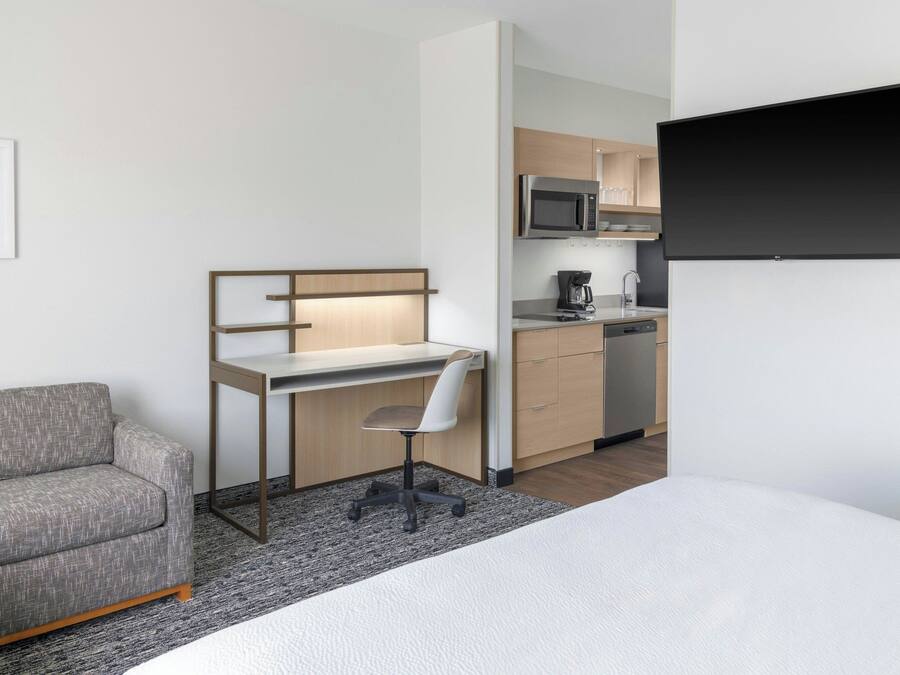 Habitación de hotel moderna con cama, sofá gris, escritorio con silla, televisor de pantalla plana y kitchenette.