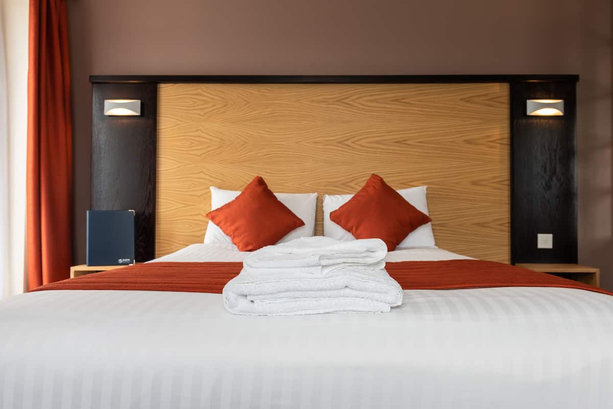 A neatly made bed with a wooden headboard, two red pillows, and a stack of white towels.