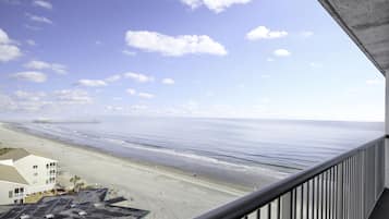 A beach view from a balcony with a railing, a pier in the distance, and a building with a flat roof.