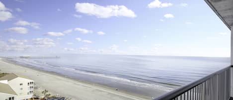A beach view from a balcony with a railing, a pier in the distance, and a building with a flat roof.