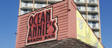 Un bar de playa con un letrero que dice "Ocean Annie's Beach Bar" y un edificio alto al fondo.