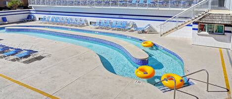 Zona de piscina con sillones de descanso, puesto de socorrista y un edificio con balcones al fondo.