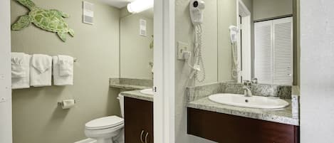 A bathroom with a green turtle wall decoration, a white toilet, a sink with a marble countertop, and a hairdryer mounted on the wall.