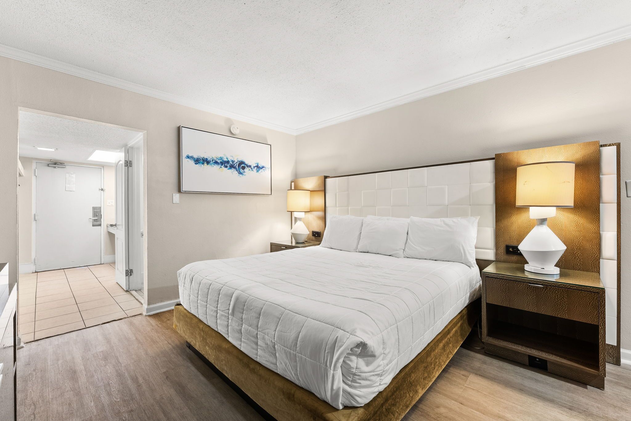 Een hotelkamer met een groot bed, nachtlampjes, een kunstwerk in een lijst aan de muur, en een deur naar een andere kamer.