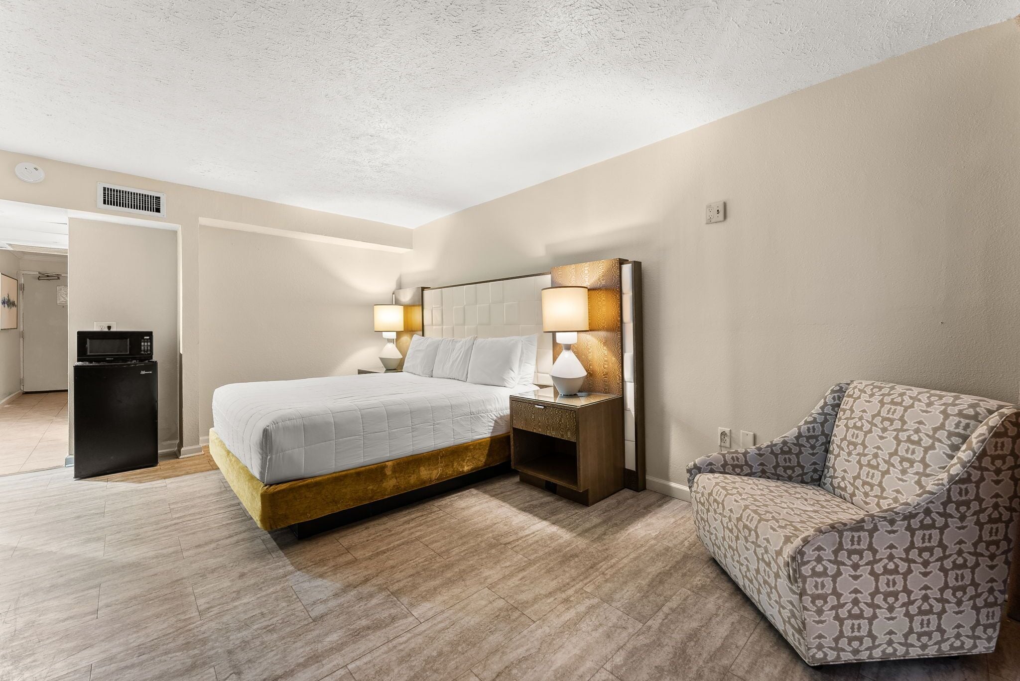 A hotel room with a bed, bedside lamps, a nightstand, a sofa, and a small refrigerator.