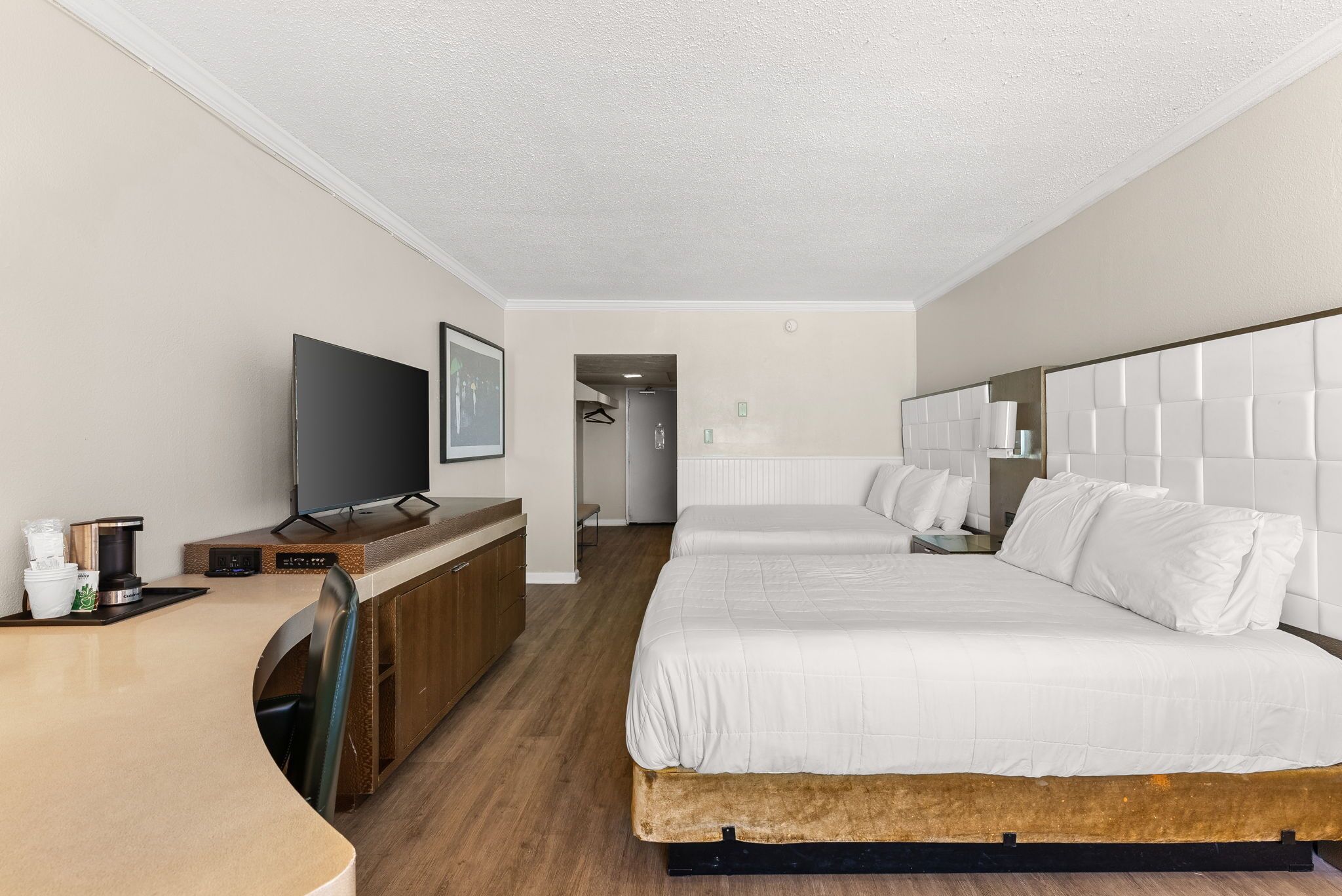 A hotel room with a bed, a TV, a desk with a coffee maker, and a view of the hallway.