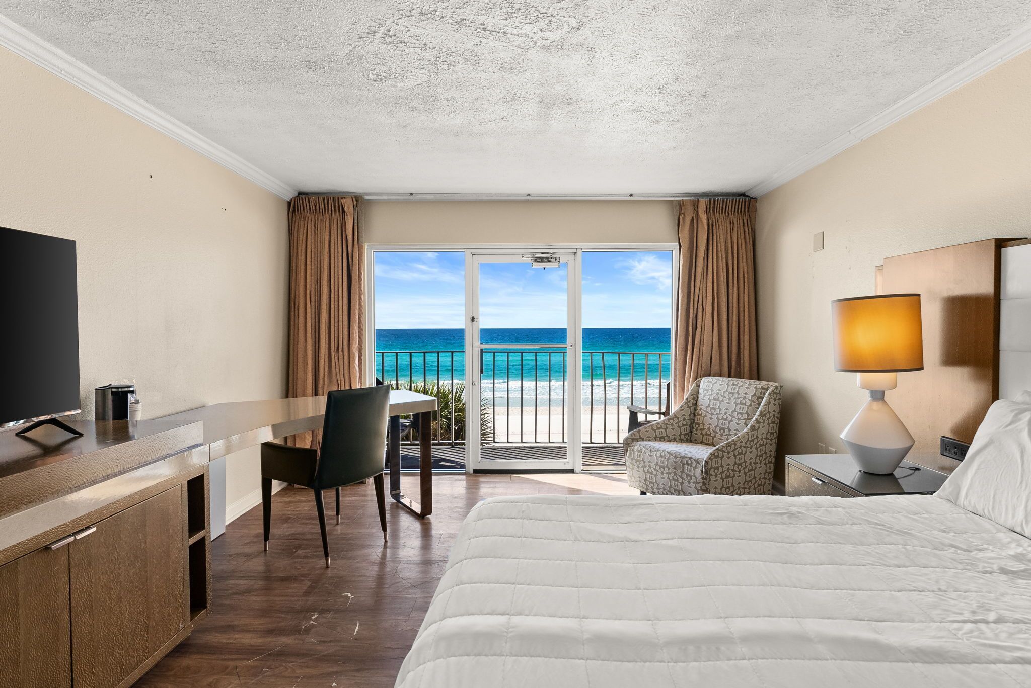 A hotel room with a bed, a desk with a TV, a chair, a lamp, and a view of the ocean.