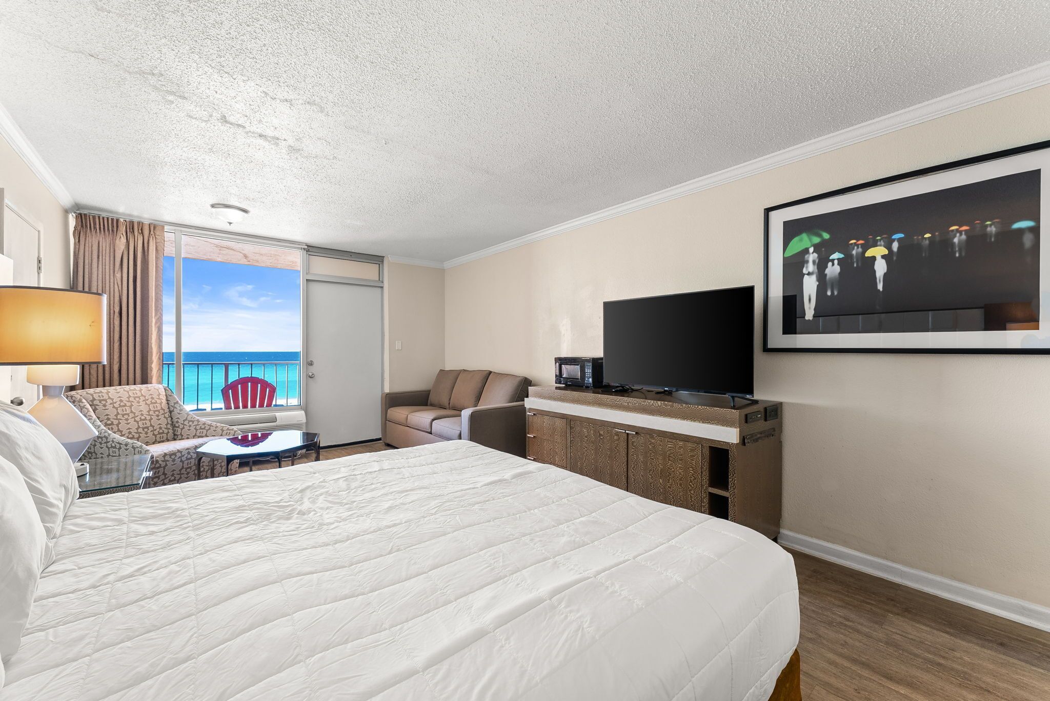 A hotel room with a large bed, a TV, a desk, a chair, a small table, and a view of the ocean.