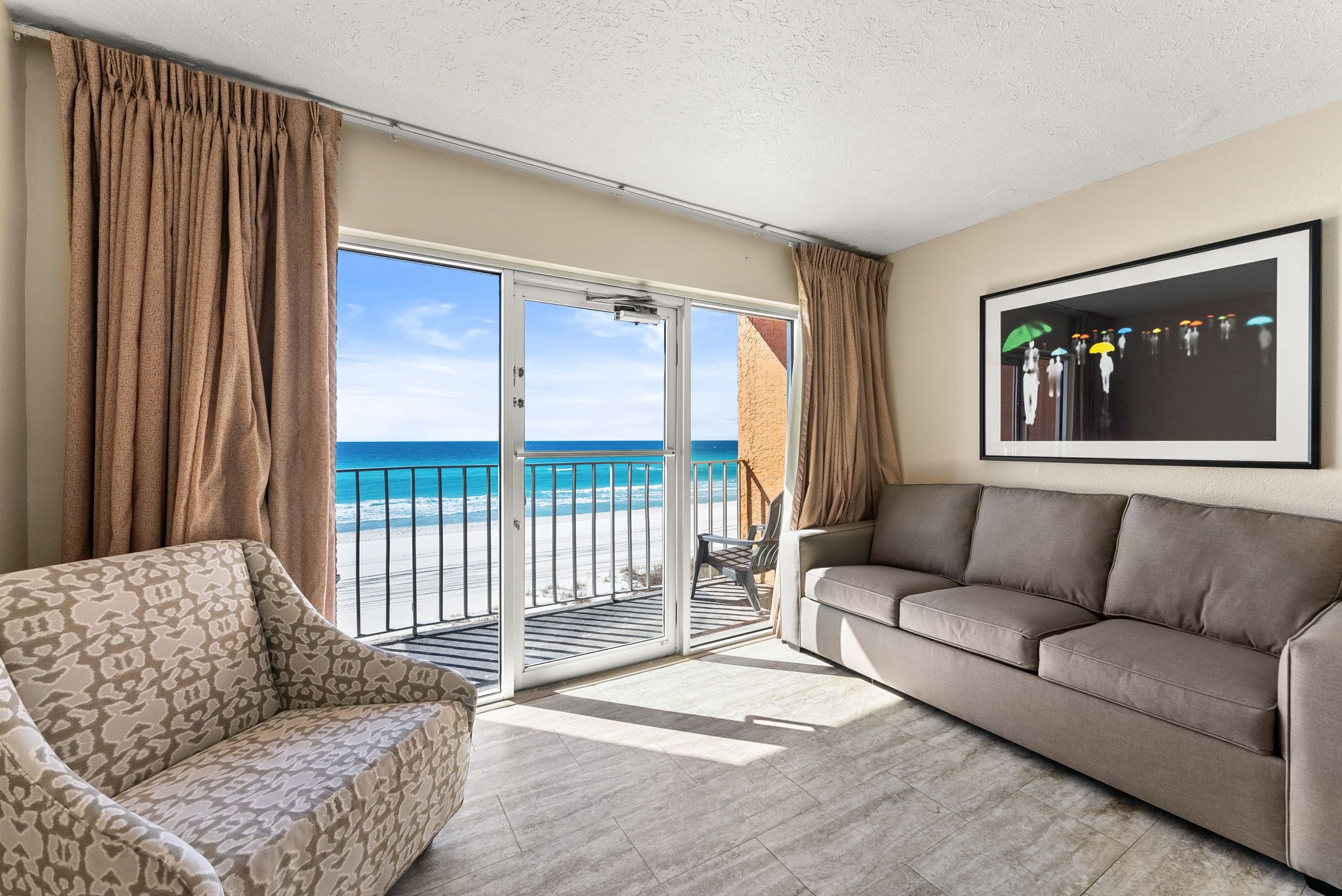 A living room with a sofa, a patterned armchair, and a view of the beach through a sliding glass door.