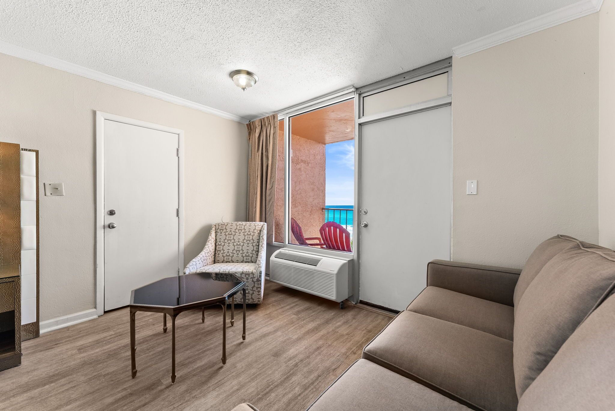 A room with a sofa, a chair, a small table, a sliding door, and a view of the outside.