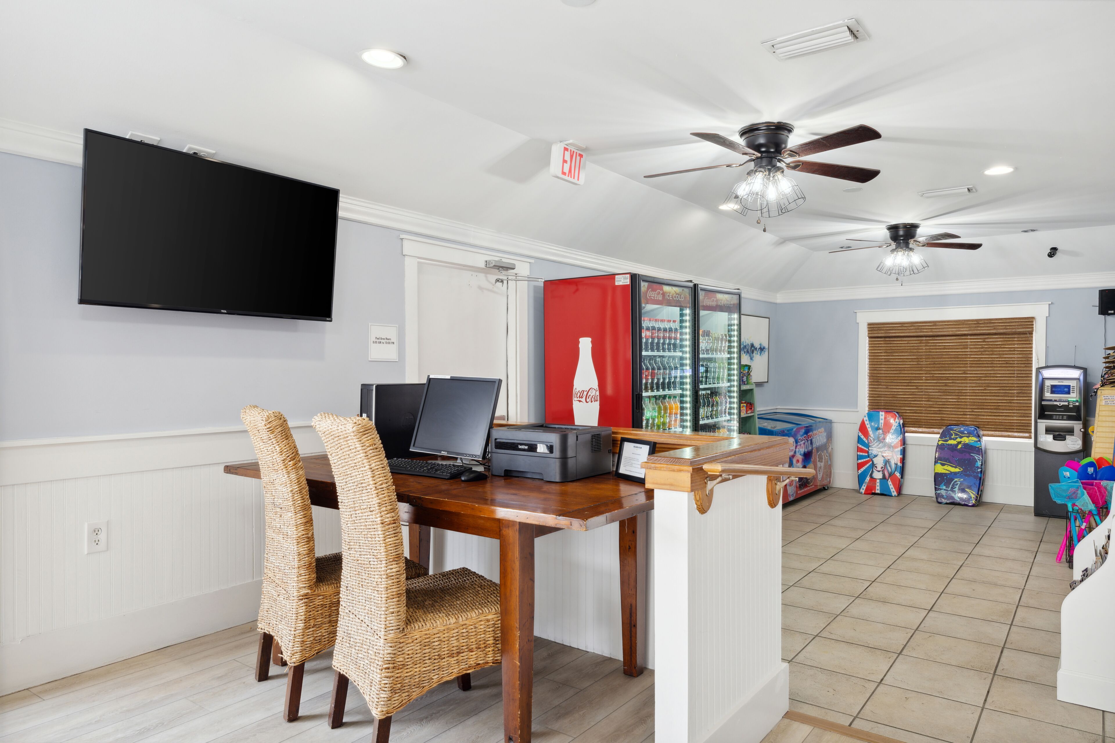 A room with a flat-screen TV, a desk with a laptop, a printer, and a Coca-Cola refrigerator. There are two ceiling fans, a window with blinds, and a door with an exit sign.