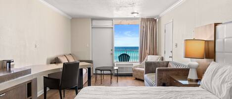 A hotel room with a bed, a desk, a chair, a small table, and a balcony with a view of the sea.