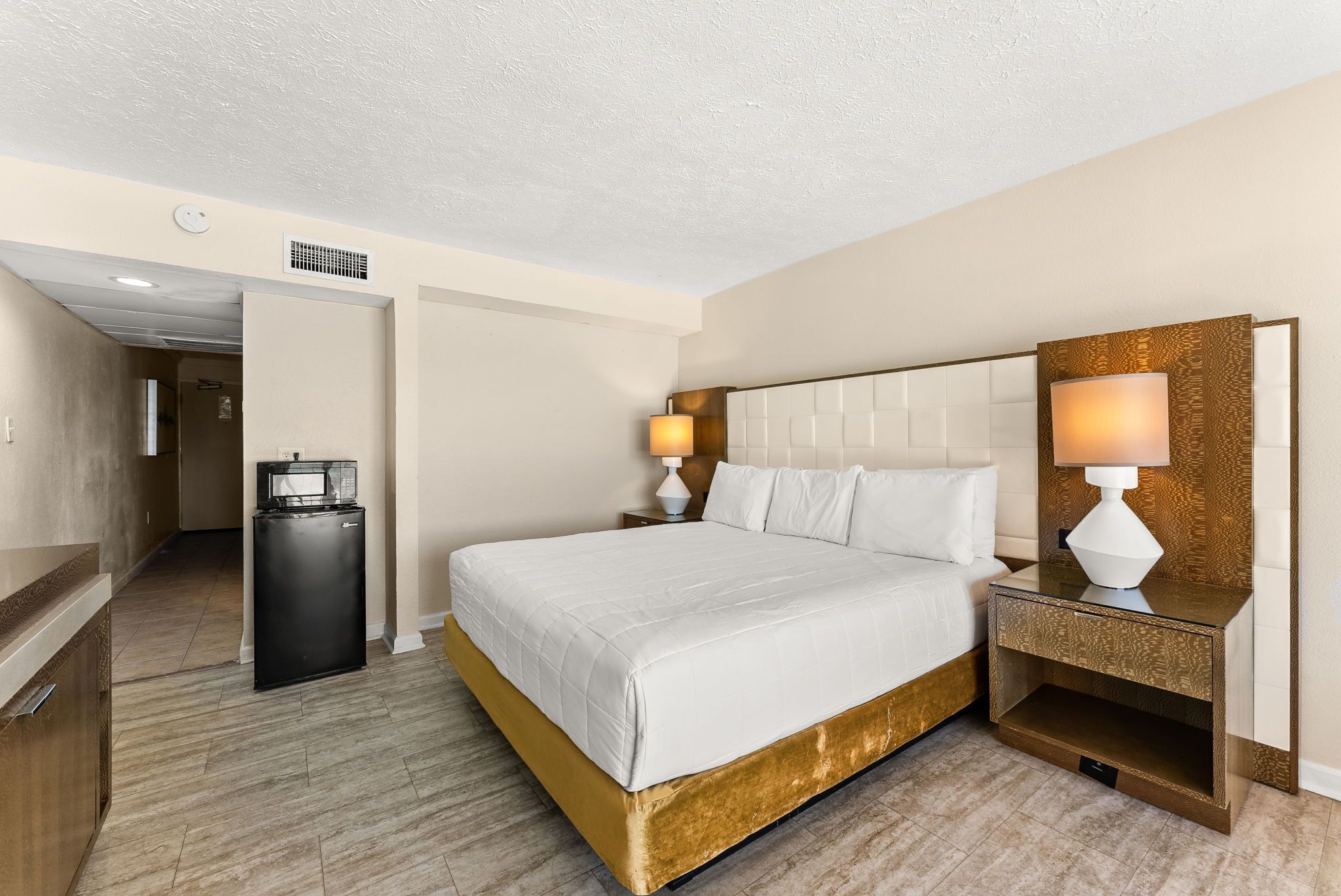 Habitación de hotel con una cama grande, dos mesitas de noche con lámparas, un minibar y una puerta que da a un pasillo.