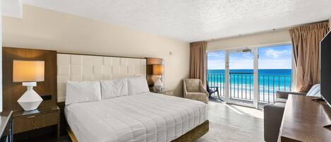 A hotel room with a large bed, a desk, a chair, and a balcony with a view of the ocean.